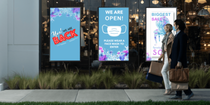 Two masked shoppers walk past three window-front retail digital signage displays on sidewalk