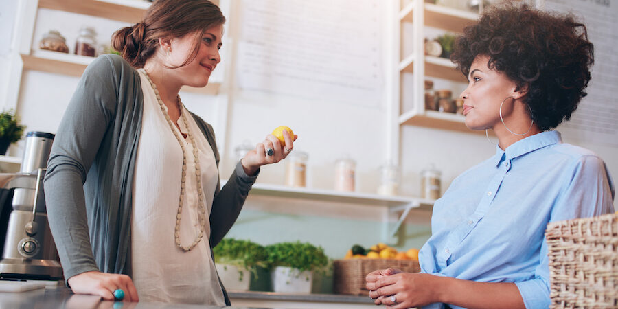 Two young female employees standing behind juice counter and walking. Juice bar owner talking with female employee.