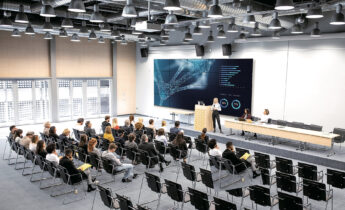 An auditorium with about two dozen seated trainees, listening to a speaker on stage in front of a large TV monitor.