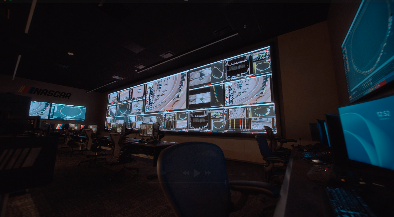 A wide-angle view from a desk in NASCAR's race control room, looking across empty office chairs toward a massive Samsung "The Wall" display filled with telemetry, race feeds, and two prominent "FS1 NASCAR" logos