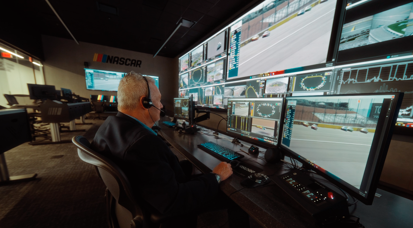 A wide-angle view from a desk in NASCAR's race control room, looking across empty office chairs toward a massive Samsung "The Wall" display filled with telemetry, race feeds, and "FS1 NASCAR" logos.