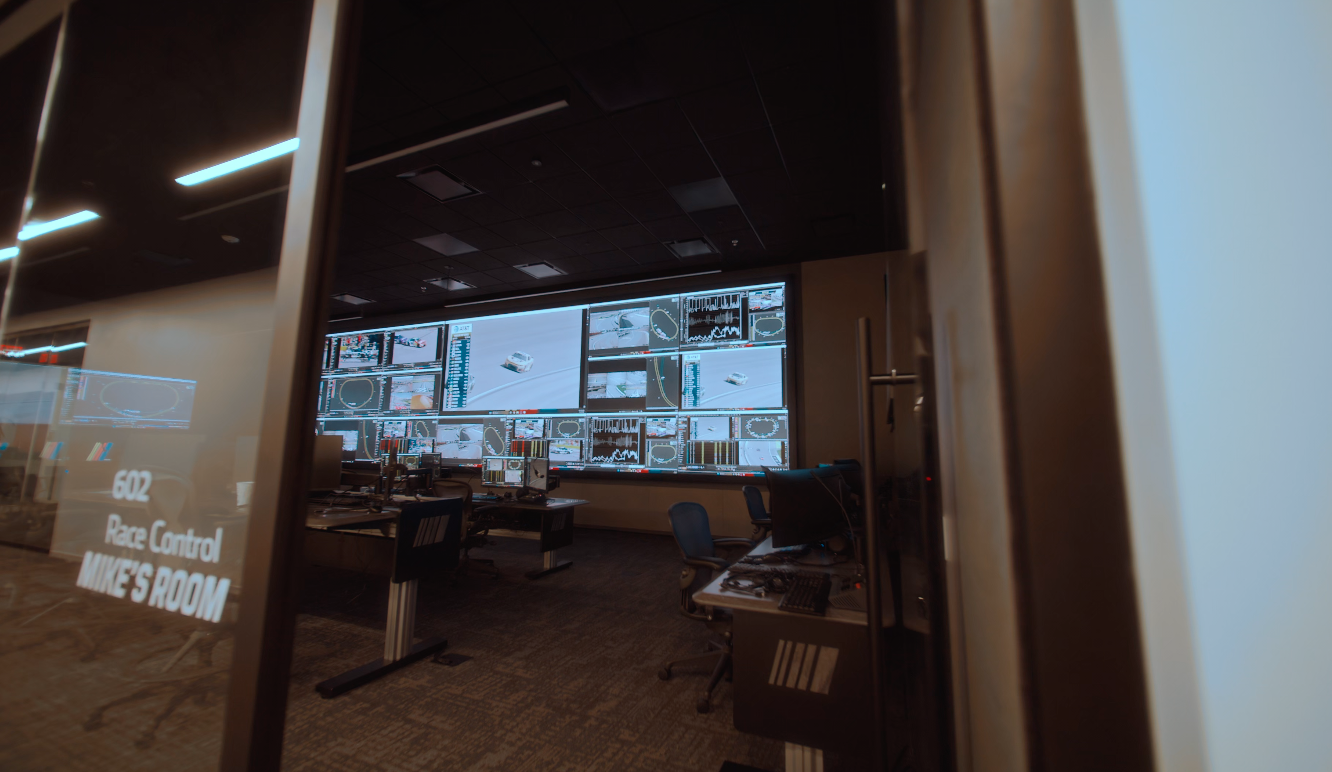 An interior photograph from a side angle inside NASCAR's remote race control facility. A man wearing a dark suit stands with his back to the camera, positioned halfway down a long dark desk lined with computer monitors and office chairs. He is looking up at a massive Samsung "The Wall" microLED display that dominates the left side of the room. The giant screen shows a mosaic of live stock car racing, multi-angle track cameras, leaderboards, and oval track telemetry. In the background, the colorful NASCAR logo is mounted on the wall next to two additional secondary displays.