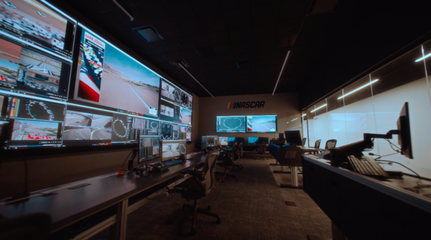 A wide-angle, side-profile view of the NASCAR Remote Race Control Room, showing a long desk lined with monitors and office chairs facing a massive Samsung "The Wall" microLED screen filled with live race feeds and track data.