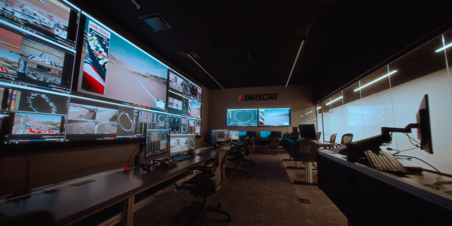 A wide-angle, side-profile view of the NASCAR Remote Race Control Room, showing a long desk lined with monitors and office chairs facing a massive Samsung "The Wall" microLED screen filled with live race feeds and track data.
