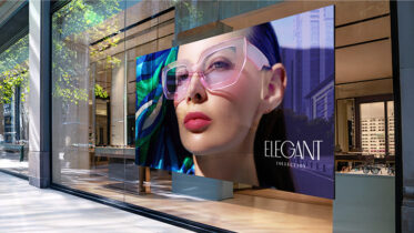 A large, vibrant Samsung indoor LED signage display installed behind a glass storefront, showcasing a fashion advertisement featuring a woman in pink glasses and the text "ELEGANT COLLECTION."