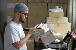 A retail or warehouse worker with a beard, glasses, and a grey beanie looks at a smartphone in his right hand while holding a small cardboard shipping box in his left hand. He is standing at a desk in a stockroom surrounded by stacked cardboard boxes, shipping labels, and shoes. A laptop, a barcode scanner, and a glass of coffee sit on the desk in front of him.