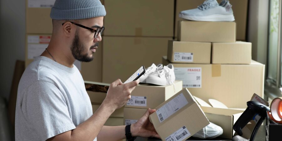 A retail or warehouse worker with a beard, glasses, and a grey beanie looks at a smartphone in his right hand while holding a small cardboard shipping box in his left hand. He is standing at a desk in a stockroom surrounded by stacked cardboard boxes, shipping labels, and shoes. A laptop, a barcode scanner, and a glass of coffee sit on the desk in front of him.