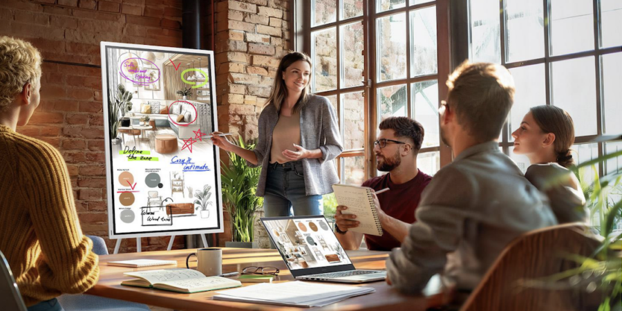 A woman stands in a sunlit, rustic office with exposed brick walls and large grid windows, leading a meeting with four colleagues seated around a wooden conference table. She points a stylus toward a large, vertically oriented digital WMFX display displaying an interior design concept, complete with sketches of a living room, color swatches, and handwritten notes like "Define the zone" and "Warm Wood tones." The seated team members—two men and two women—are looking toward her attentively. On the table in the foreground are an open laptop showing the same design concept, notebooks, a coffee mug, and various papers.